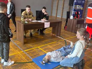 Uczennice udzielają pierwszej pomocy osobie poszkodowanej, ich zadaniu przyglądają się strażacy